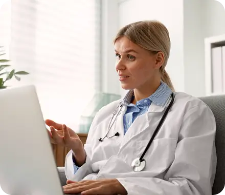A woman in a doctor's coat sits at a desk, focused on her laptop screen.